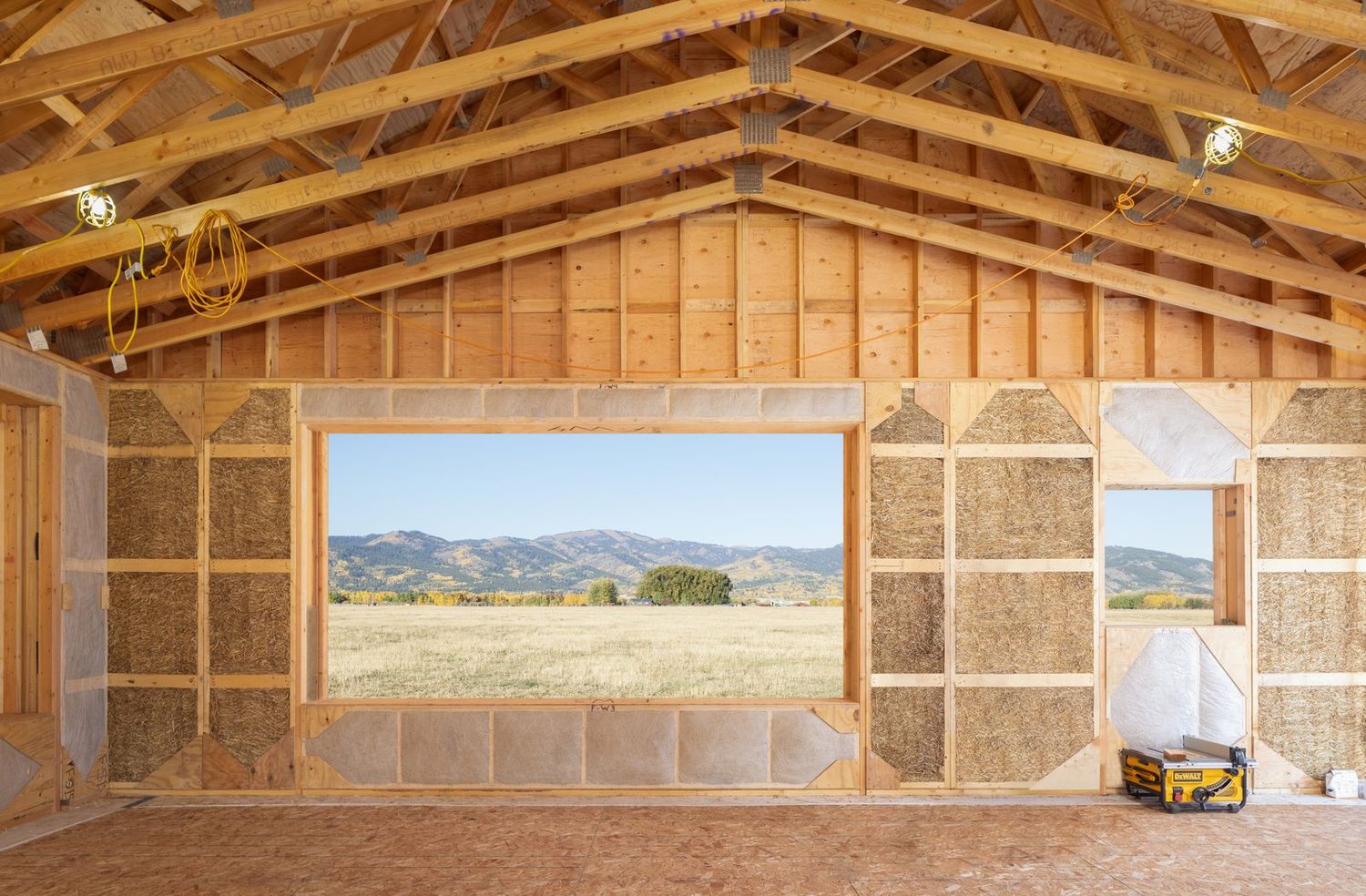 Inside a house under construction: straw panel walls, exposed roof framing, and a wide window opening onto a harvested field