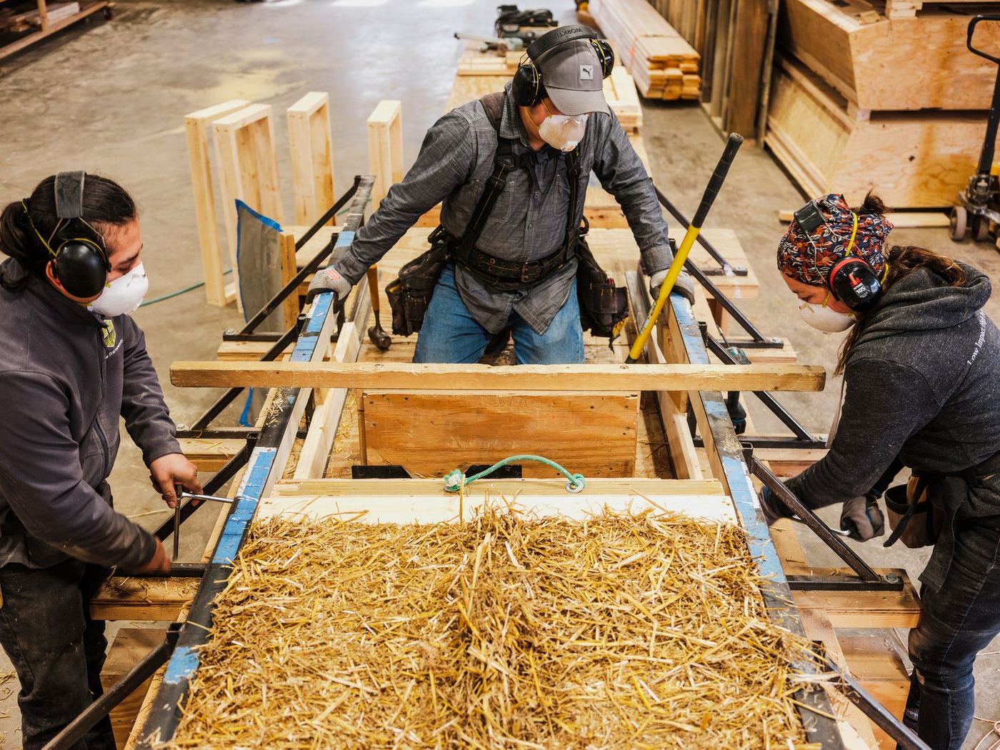 Three workers in ear defenders and dust masks pressing straw into a timber panel frame